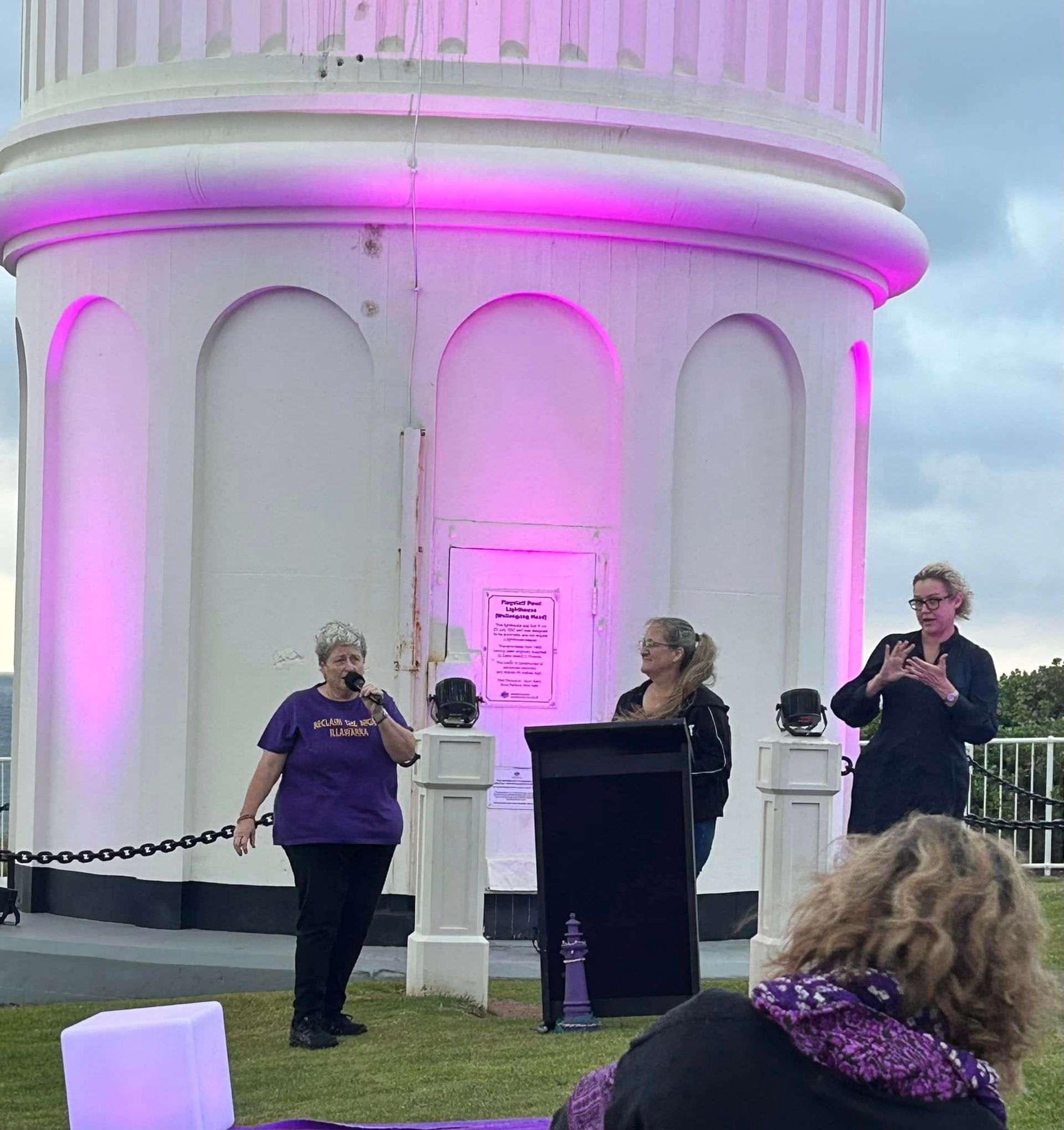 People speaking at a podium in front of a lighthouse lit with pink, outdoors.