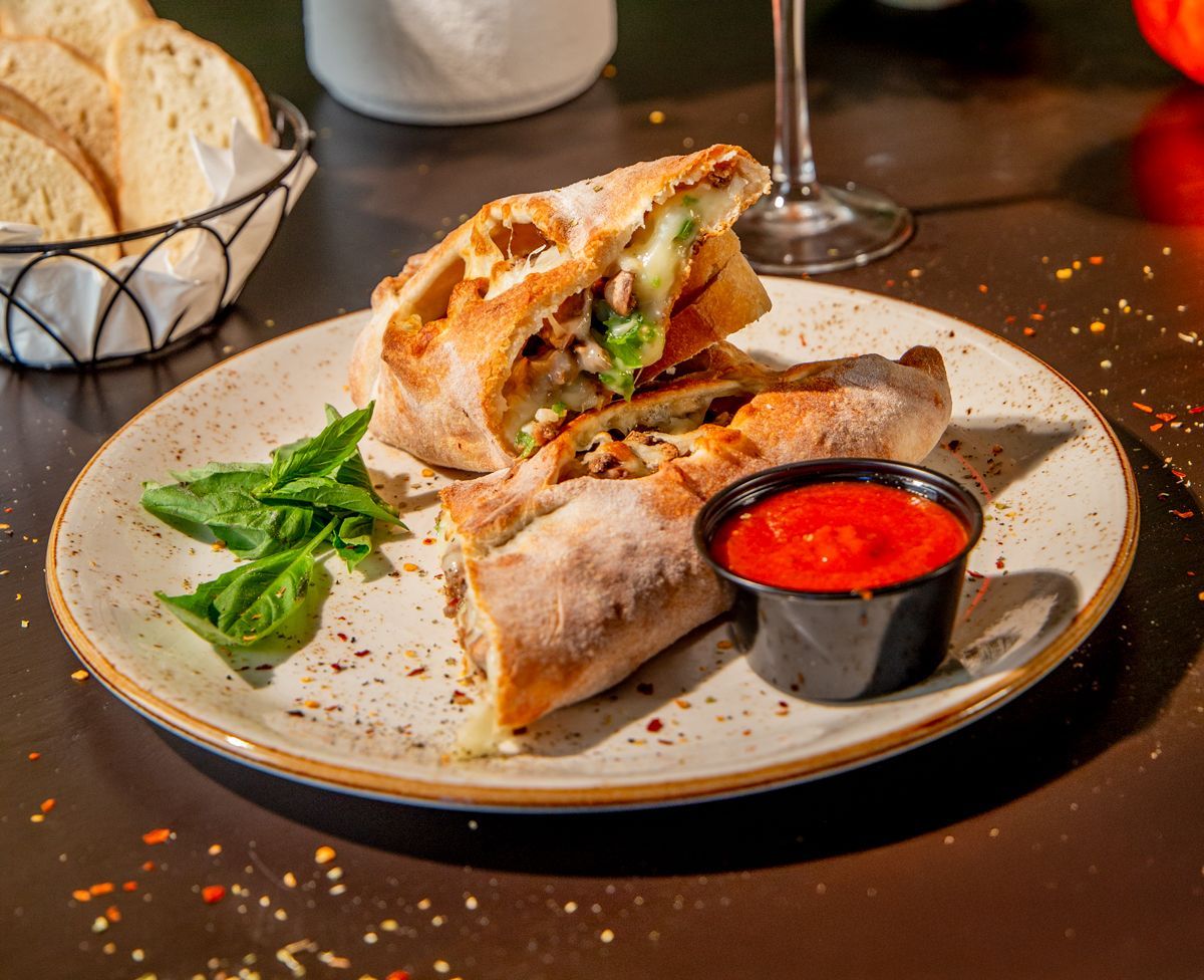 Calzone cut open on a plate with dipping sauce and basil. Bread basket in the background.