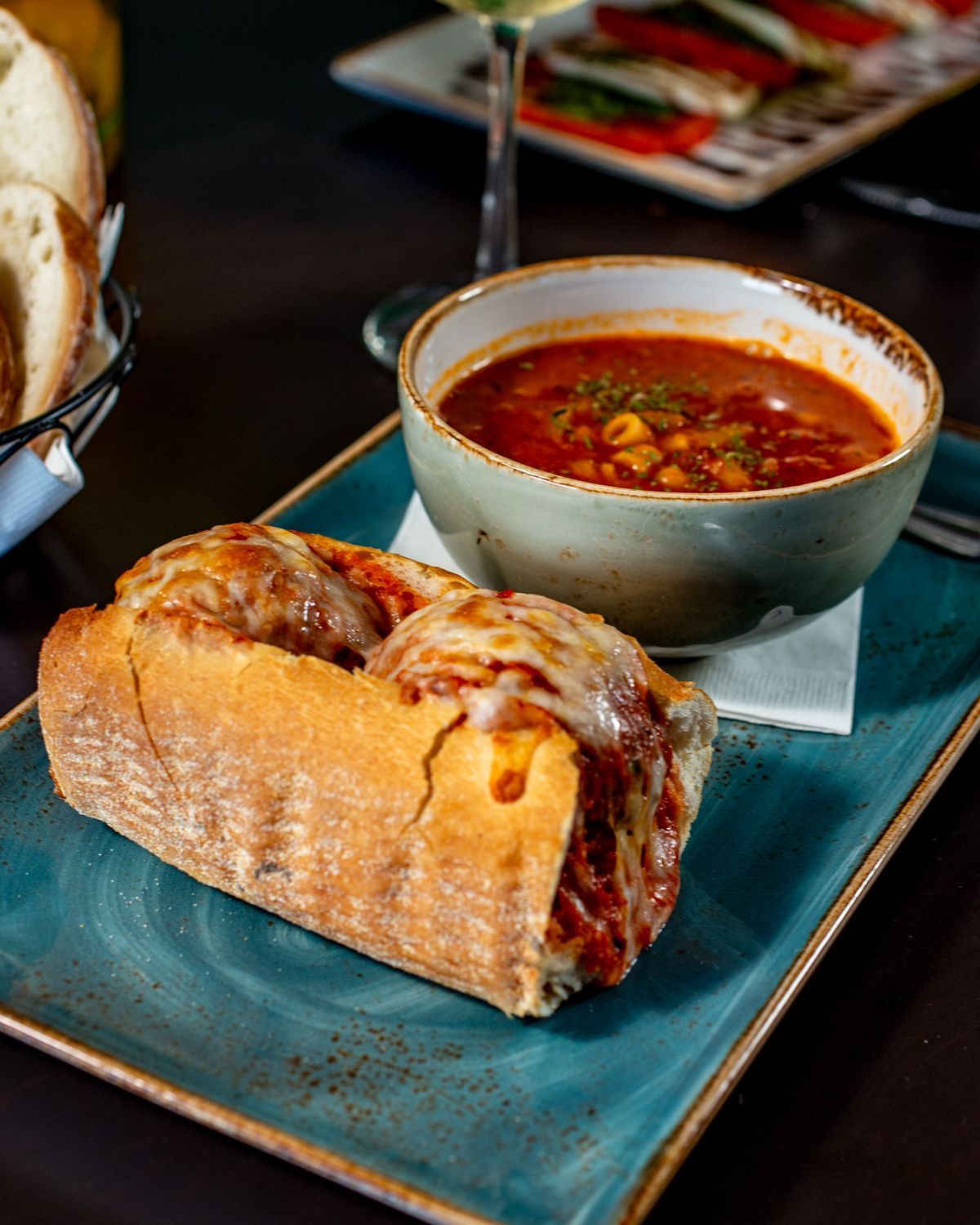 Meatball sub and soup on a blue plate, with bread in the background.
