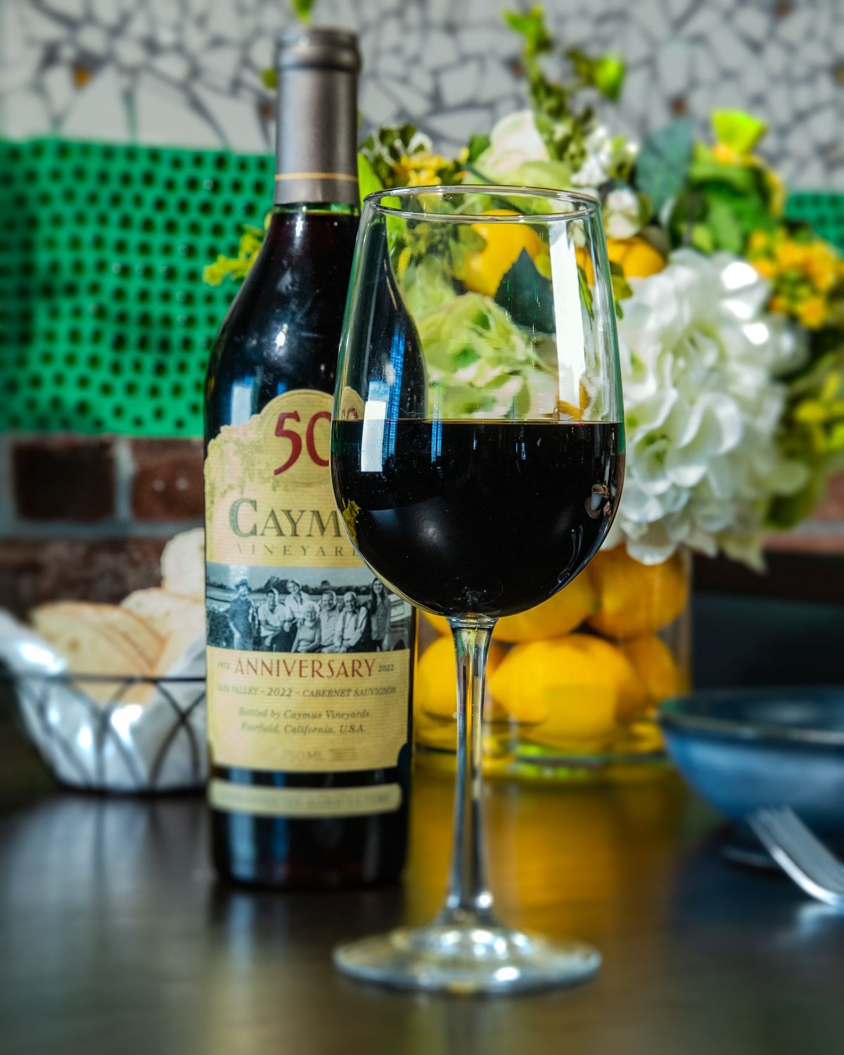 Wine bottle and glass filled with red wine, with a floral arrangement in the background.