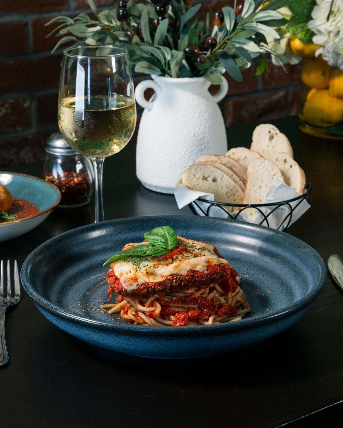 Plate of lasagna, spaghetti, and bread with a glass of wine on a table with a decorative vase and brick wall.