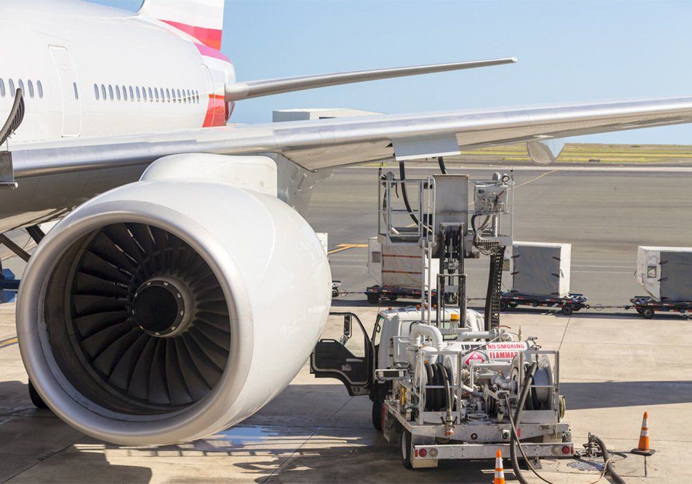 Truck parked under an aircraft wing refuelling the tanks with aviation fuel through an open flap with a turbo engine in the foreground during servicing at the airport
