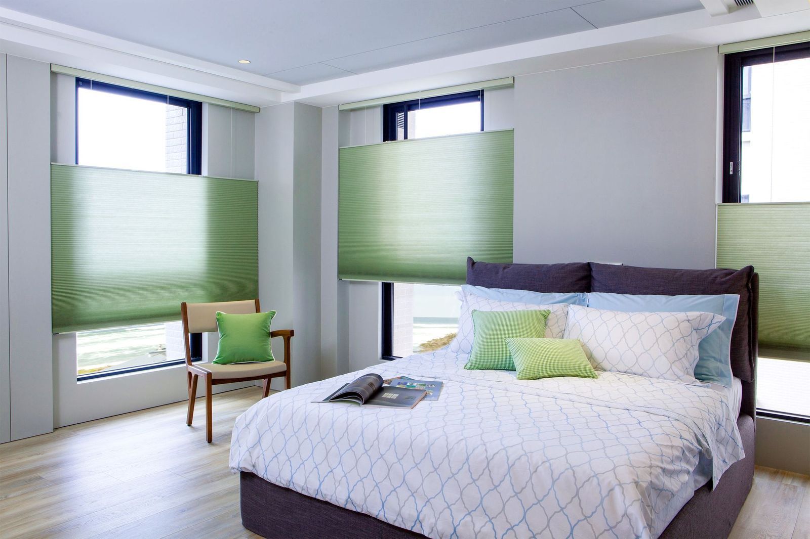 A bedroom with a bed featuring patterned bedding, light green pleated window shades, and a wooden chair in a modern space.