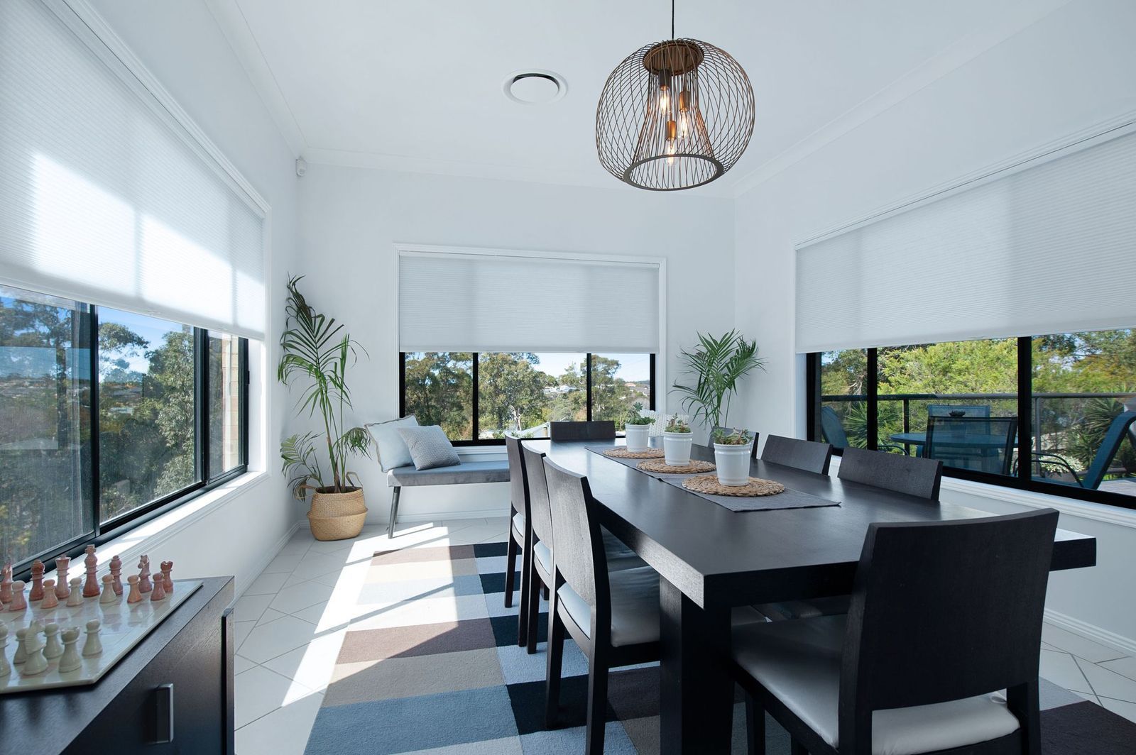 A modern, sunlit dining room features a dark table with chairs, a decorative chandelier, and large windows with blinds.