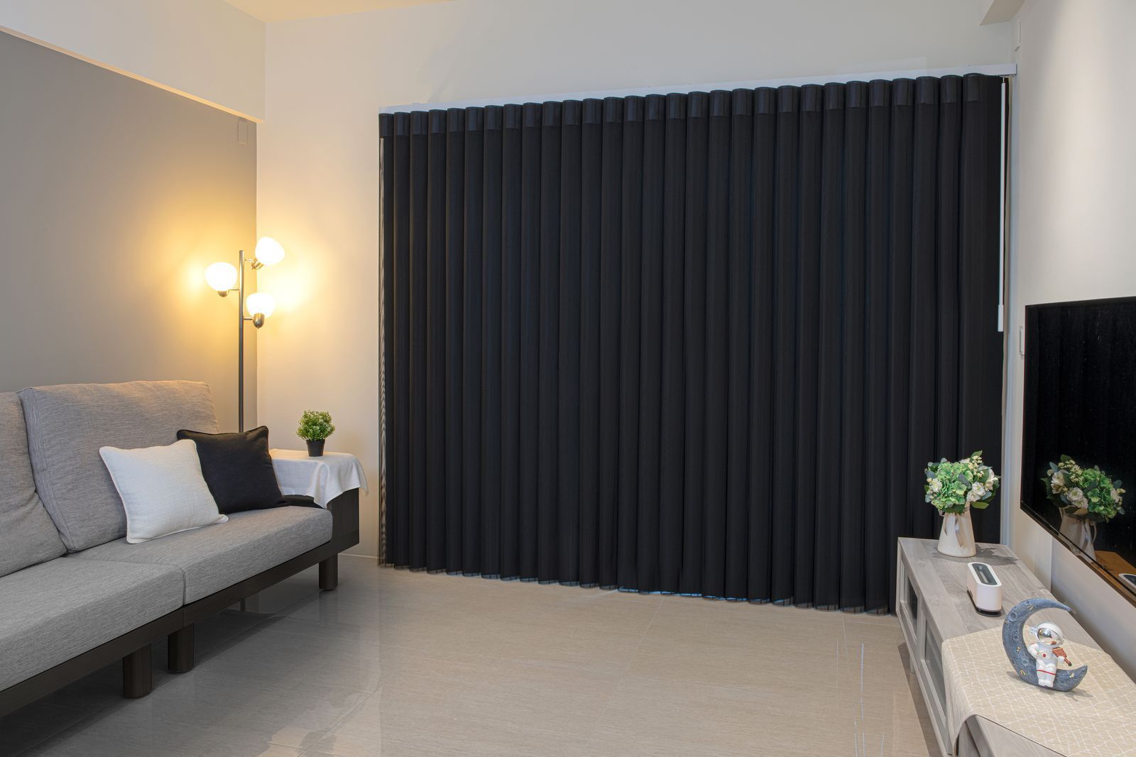 A modern living room with a gray sofa, a floor lamp, a television, and floor-to-ceiling dark gray vertical blinds.