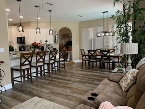 An open-concept dining and kitchen area with light walls, wood-look flooring, bar stools, and a brown sofa in the foreground.