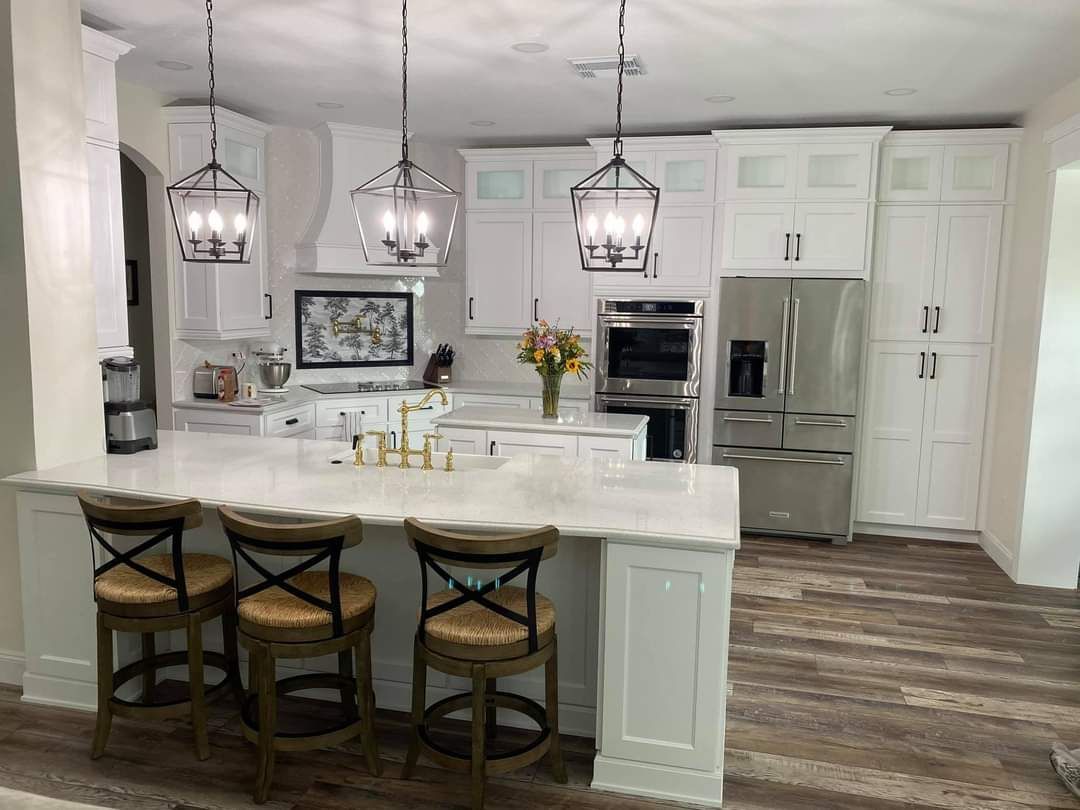 A modern white kitchen with a marble-topped island, three wooden stools, pendant lights, and stainless steel appliances.