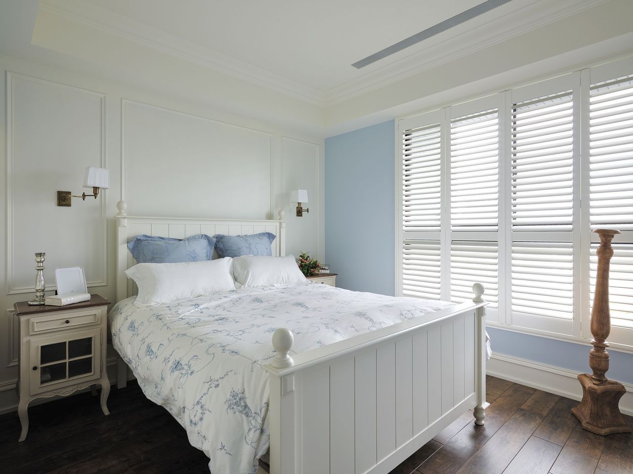 A bedroom featuring a white bed with floral bedding, a white nightstand, blue accent walls, and white window shutters.