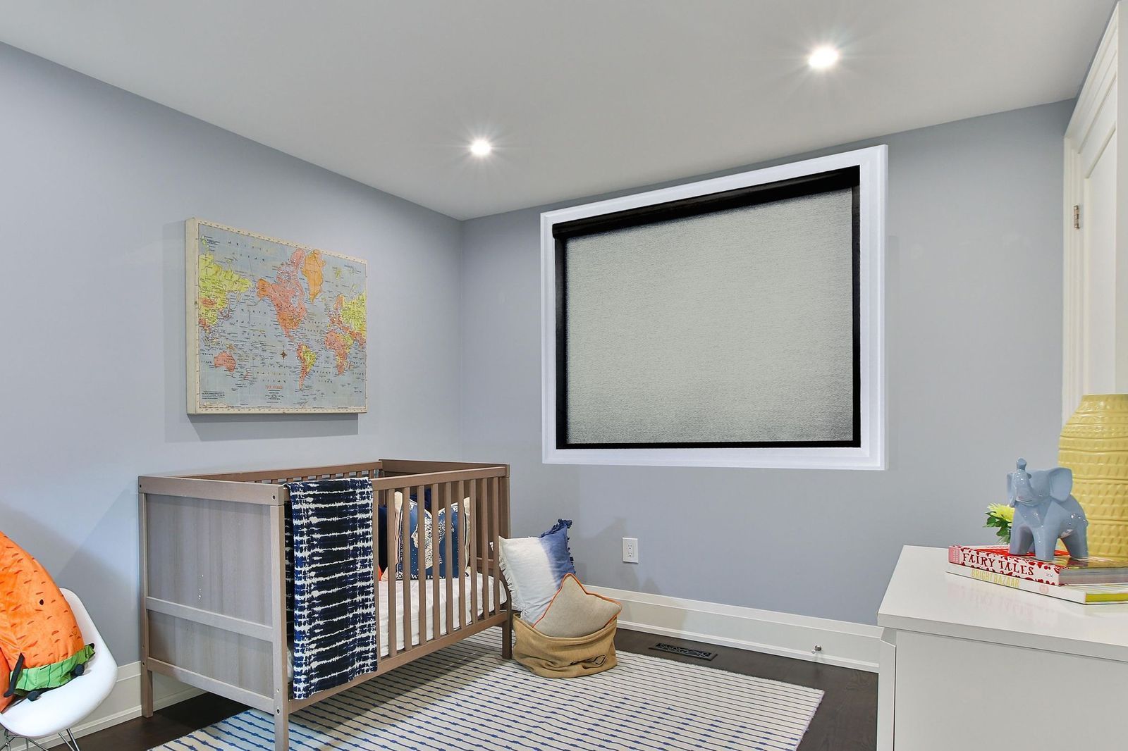 A nursery with a wooden crib, a world map wall art, and a large window with a dark roller shade against light blue walls.