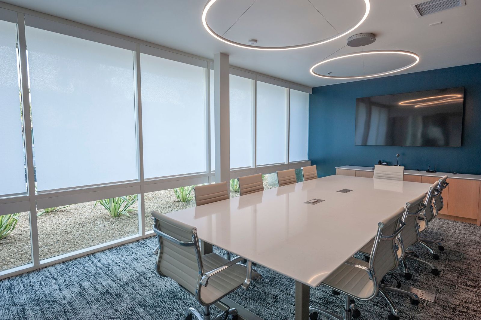 Modern conference room with a white table, chairs, large windows with shades, and a wall-mounted TV on a teal wall.