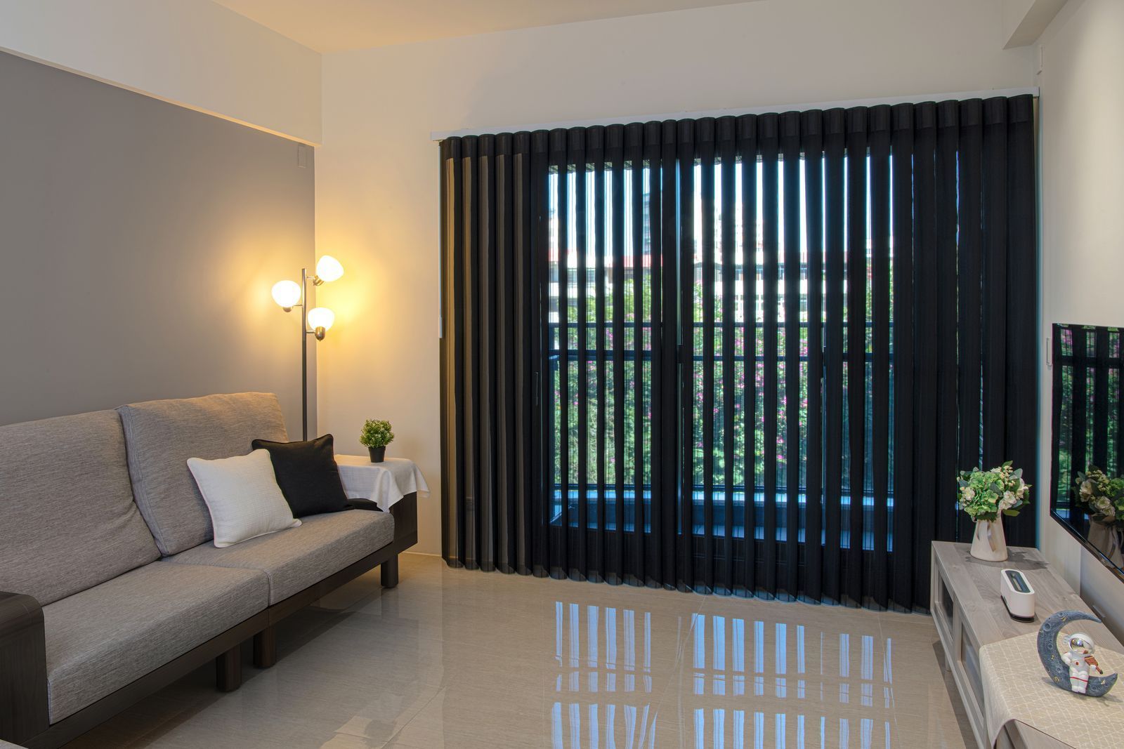 A modern living room with a grey sofa, black vertical blinds, a floor lamp, and a TV stand on a light-colored tile floor.