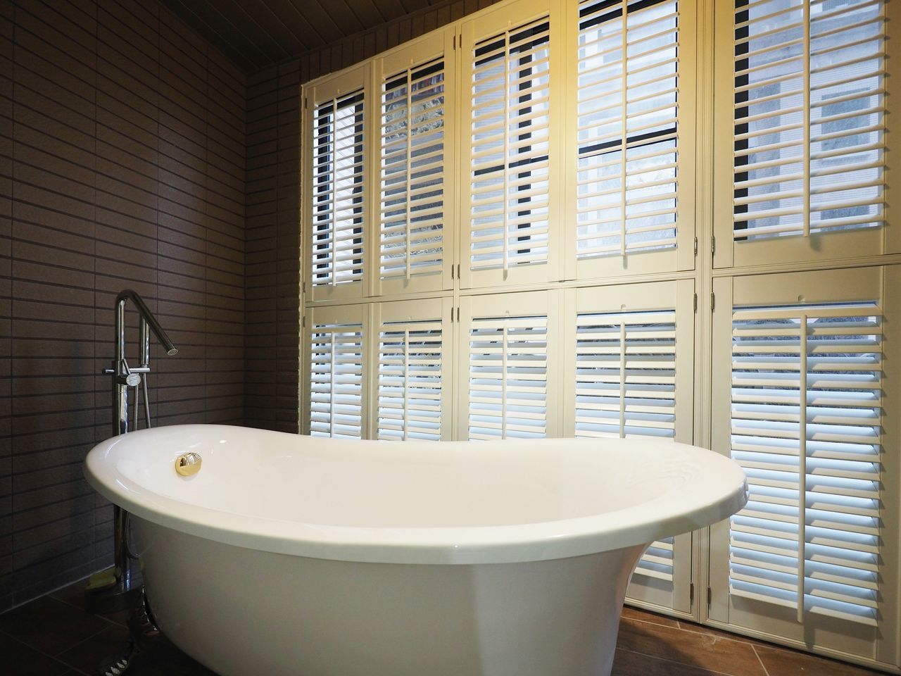 A modern white freestanding bathtub sits against a wall of dark horizontal slats and large cream-colored shutters.