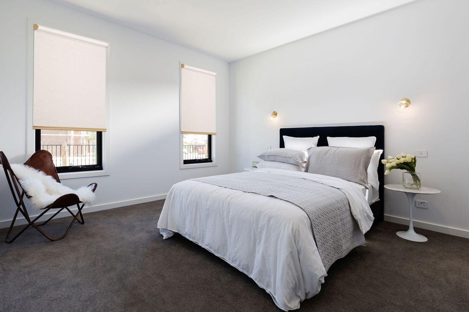 A modern bedroom with a queen bed, dark carpet, white walls, two windows with roller blinds, and a butterfly chair.