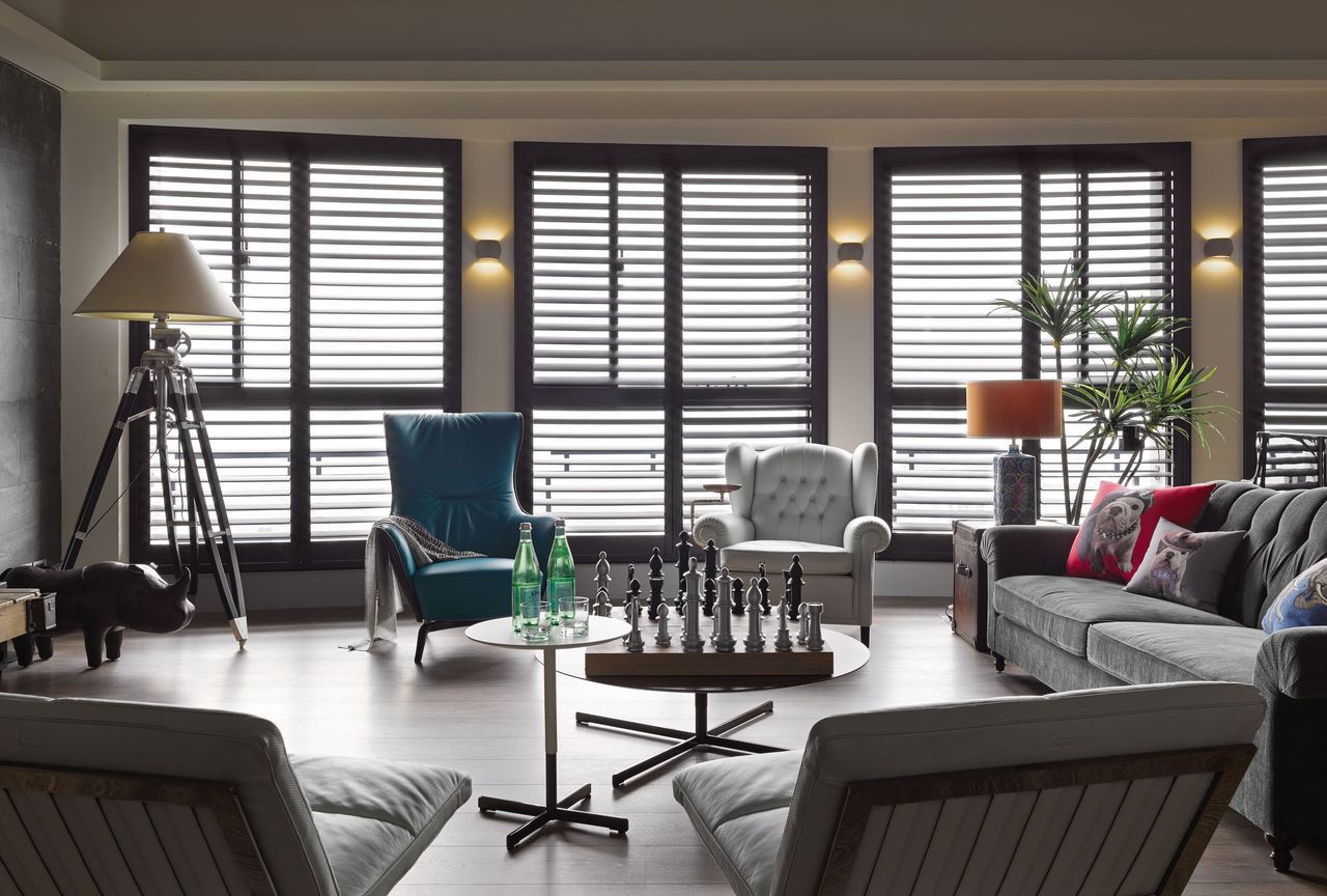 A modern living room with a sofa, armchairs, a chess set on a table, and windows with horizontal blinds.