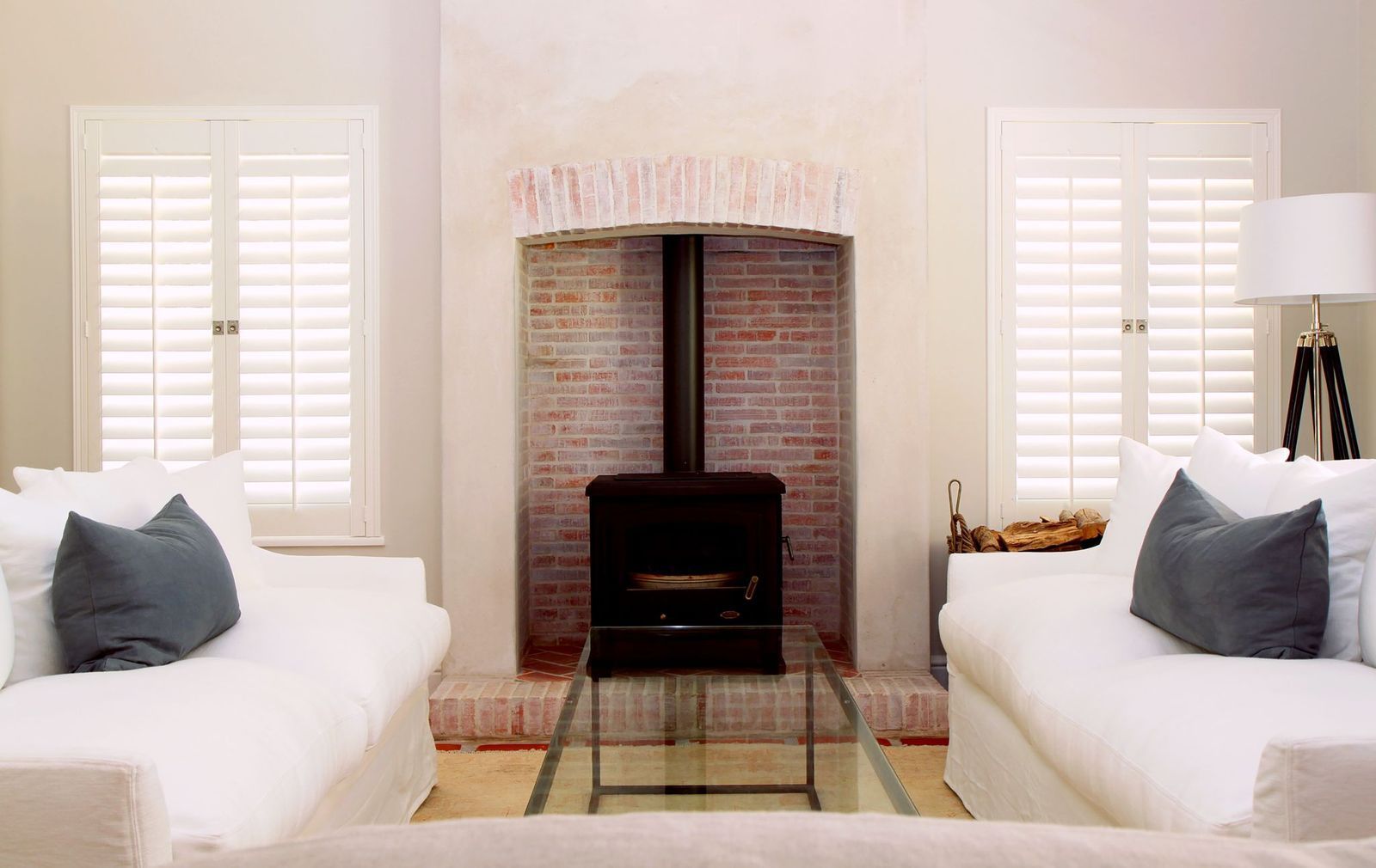A symmetrical living room with white sofas, gray pillows, white shutters, and a wood stove in a brick fireplace.