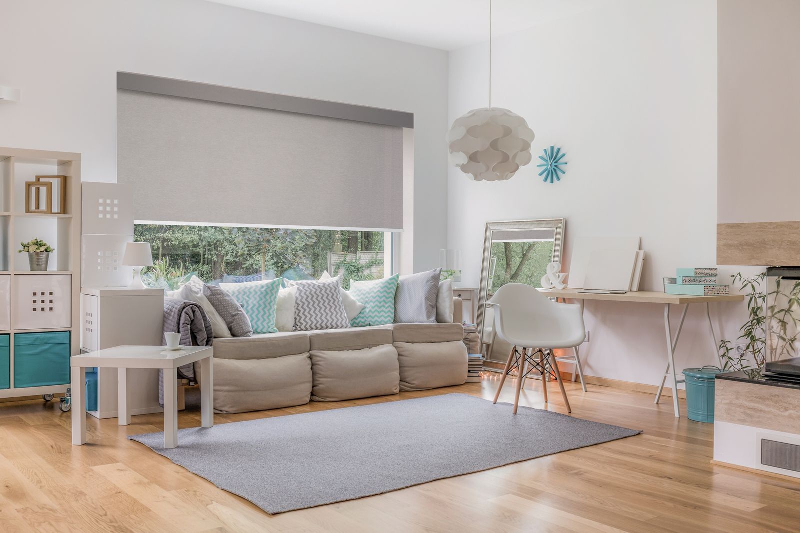 A light-filled living room with a beige sofa, blue accents, a gray rug, a desk, and a large window with a roller shade.