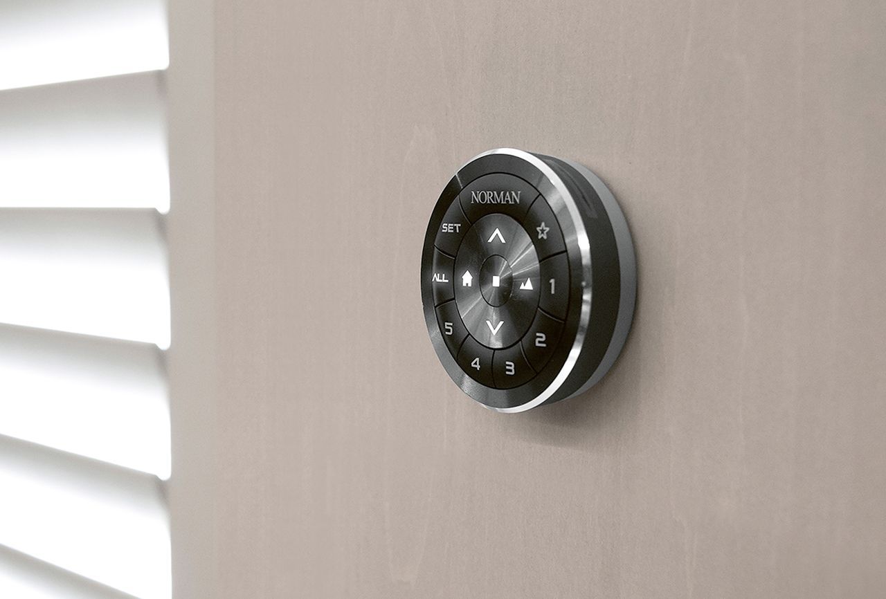 A circular, metallic, wall-mounted smart shade remote with touch controls, positioned next to white window blinds.