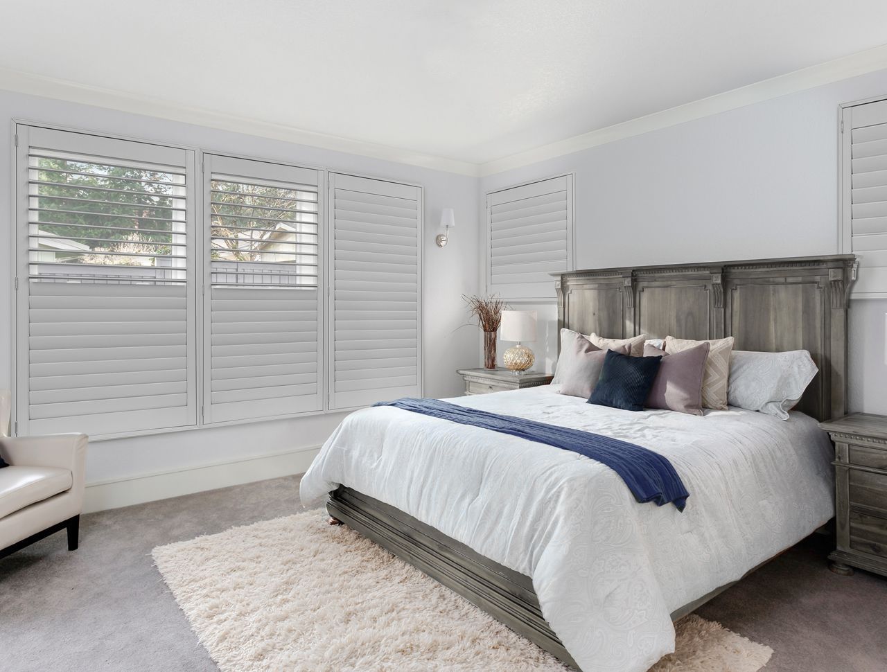 A modern, neutral-toned bedroom featuring a grey wooden bed, white plantation shutters, and a plush cream rug.