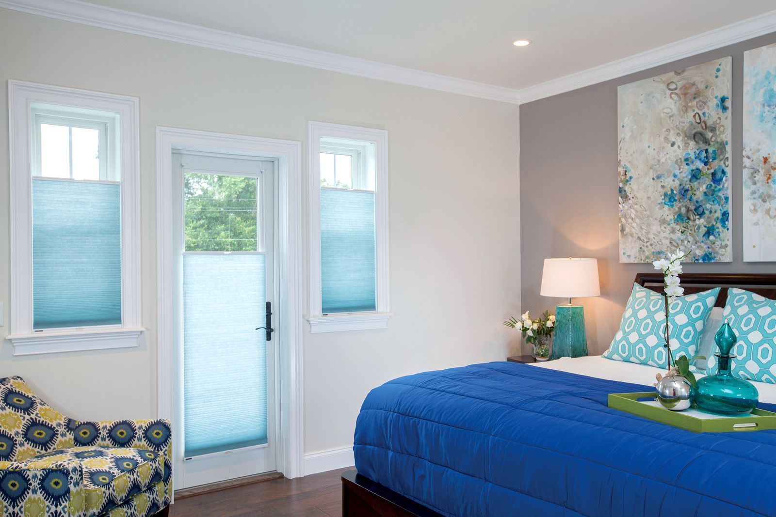 A blue-themed bedroom with two windows and a glass door featuring teal shades, a blue bedspread, and abstract wall art.