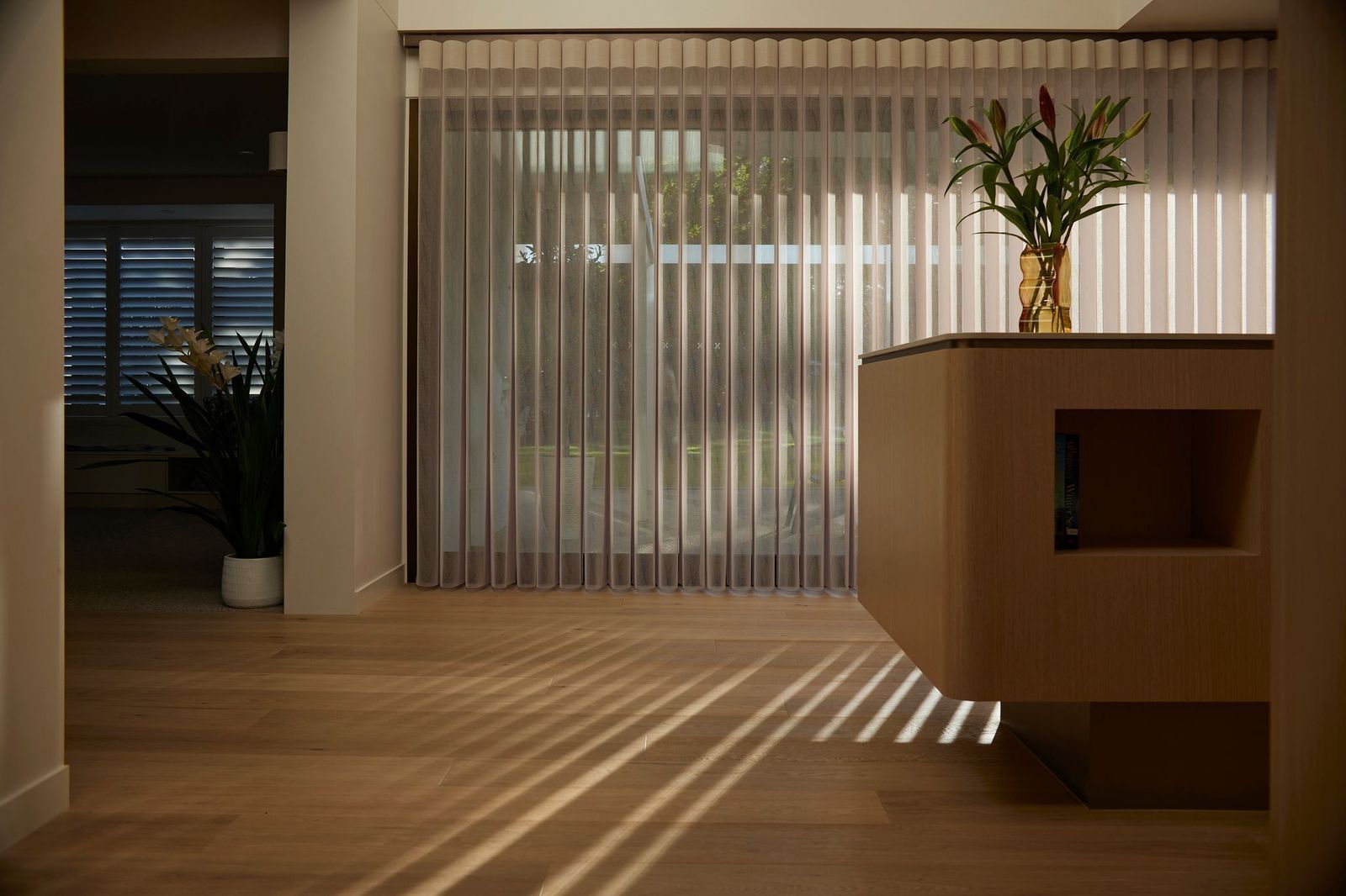 Sunlight streams through vertical blinds into a modern room, casting long shadows across a light wood floor.