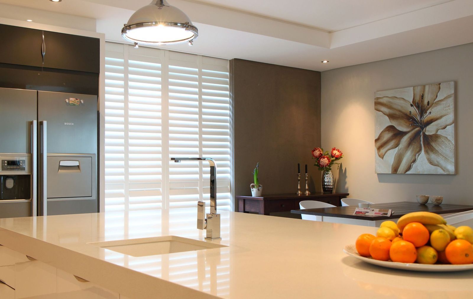 A modern kitchen with a large white island, stainless steel refrigerator, plantation shutters, and a fruit bowl.