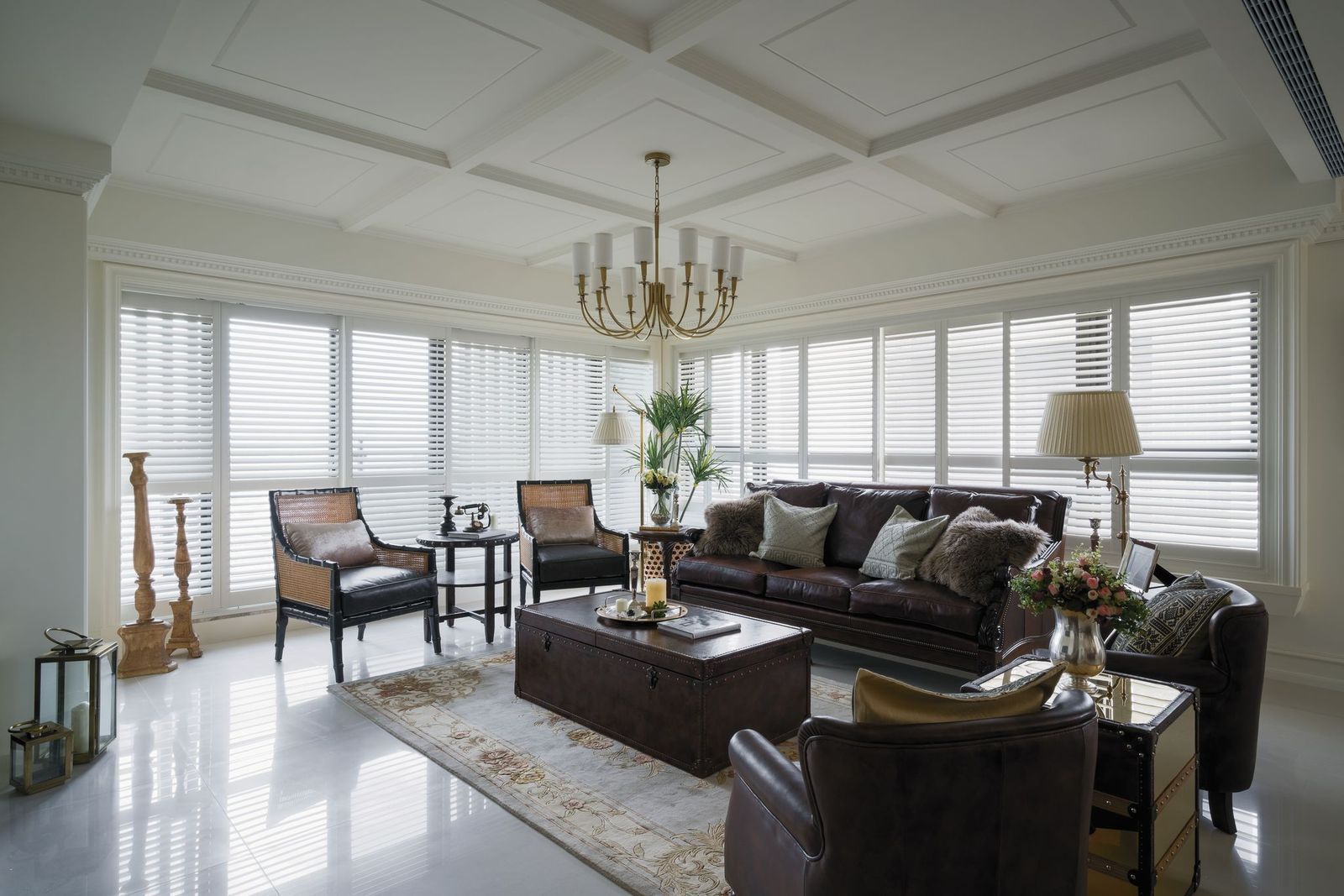 A bright, elegant living room featuring dark leather furniture, a central coffee table, and large windows with white blinds.