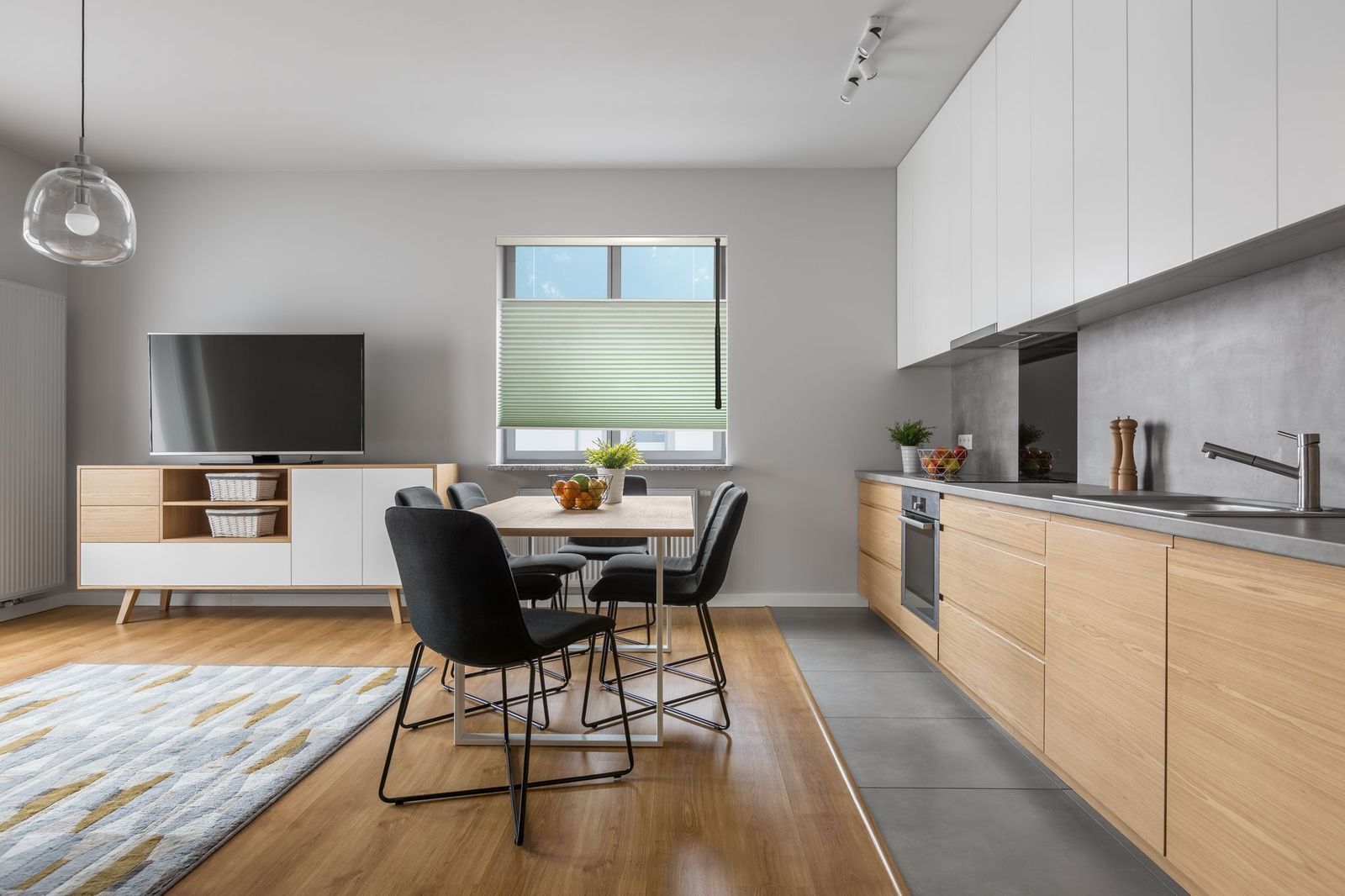 A modern open-plan kitchen and dining area with light wood cabinets, black chairs, and a television on a side console.