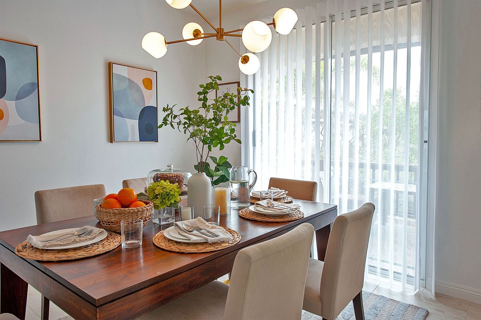 A dining room with a wooden table set for dinner, featuring a central floral arrangement, modern chandelier, and wall art.