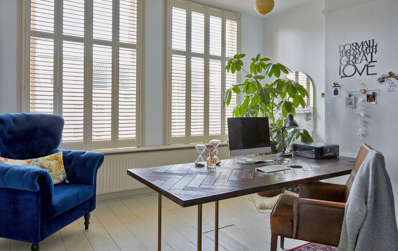 Home office featuring a dark wooden desk, blue velvet armchair, brown leather chair, and large windows with shutters.