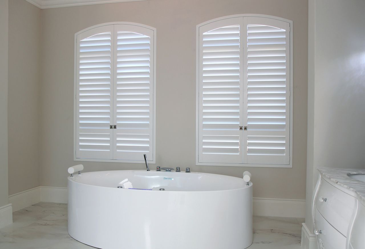 A white oval bathtub positioned in a bathroom with beige walls and two arched windows covered by white plantation shutters.