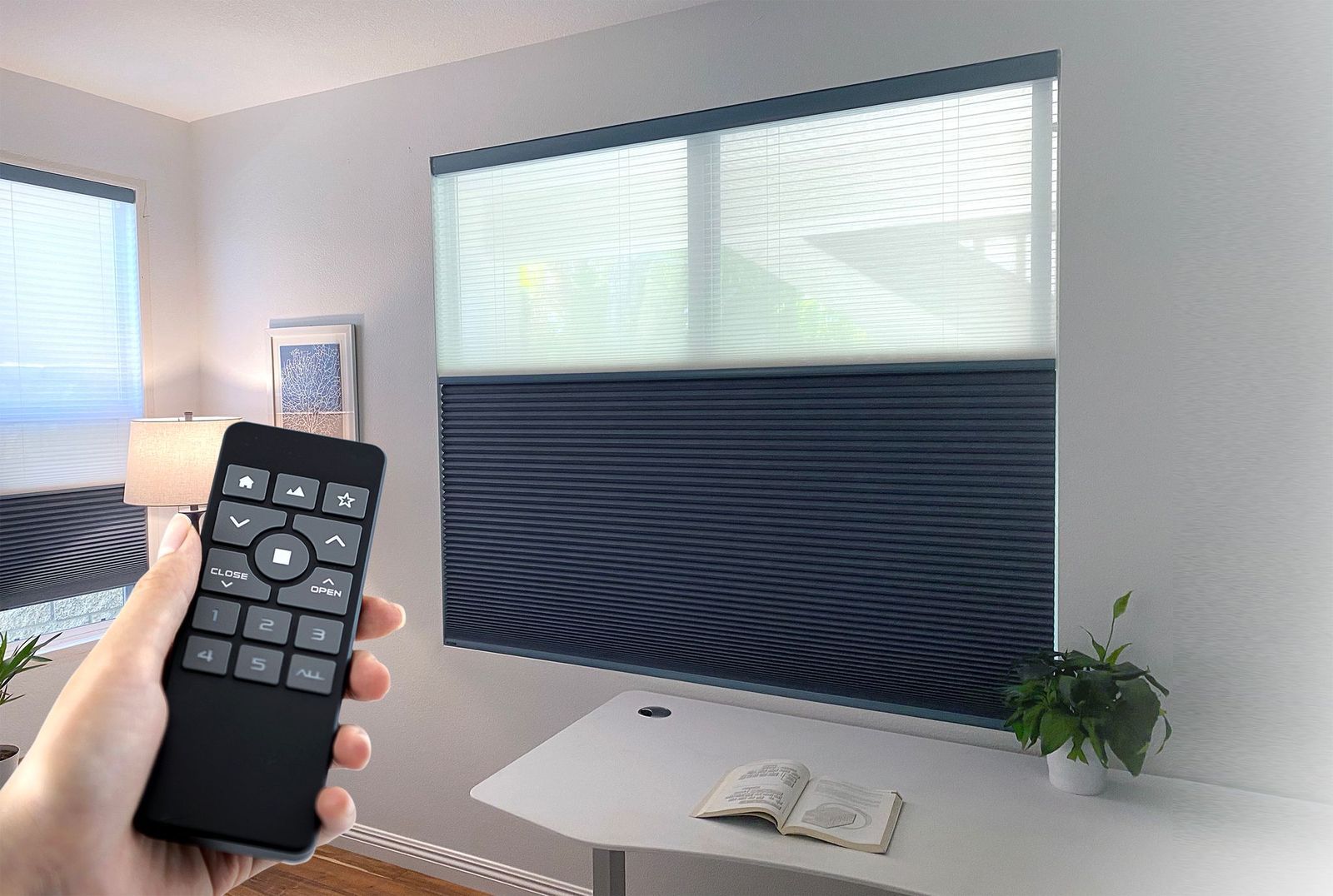 A hand holds a remote control aimed at a dual-shade window blind with white top and dark bottom sections in a room.