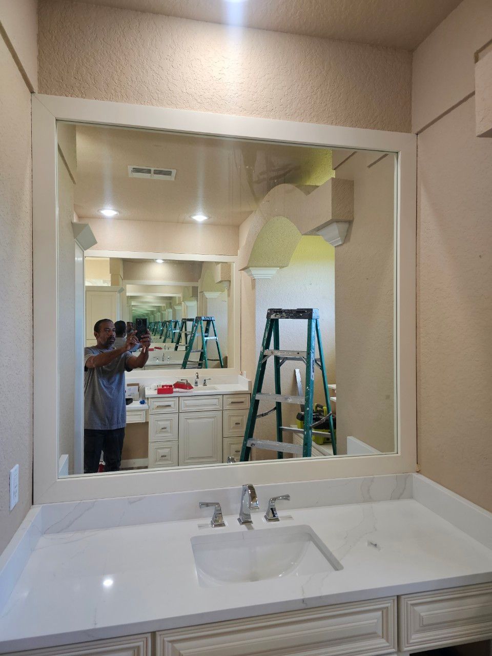A person stands in front of a bathroom vanity with a large white-framed mirror, reflecting a ladder in the room.