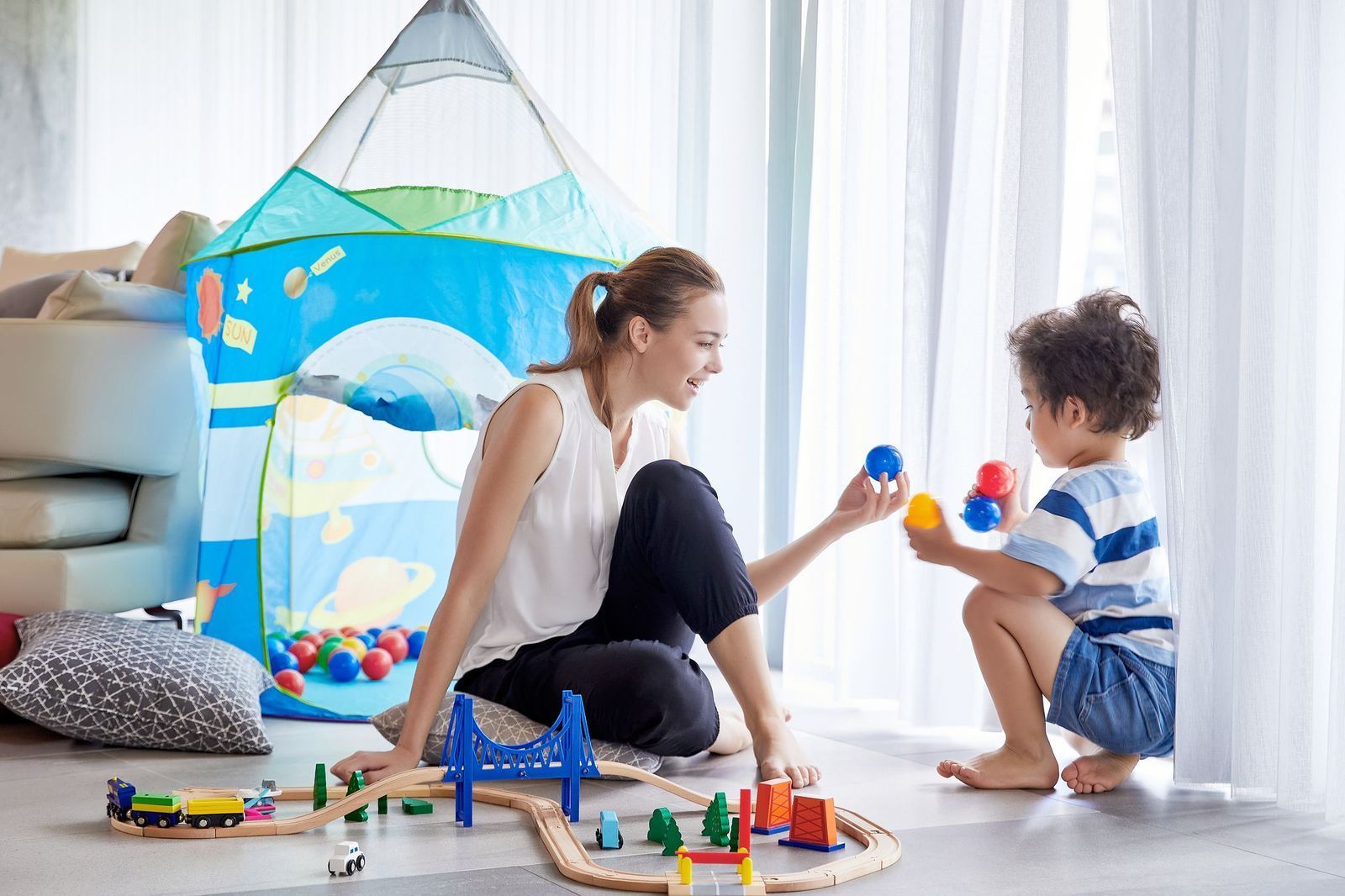 A person and a child play with colorful balls and a wooden train set on the floor in front of a blue play tent.