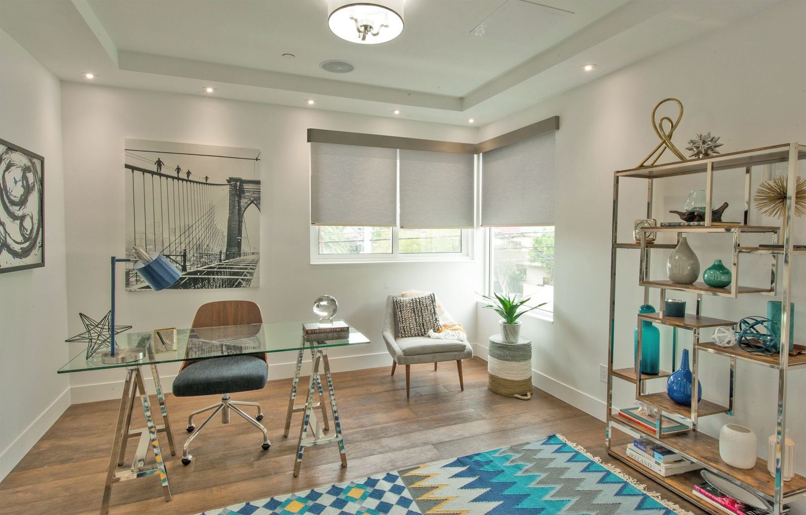 A modern home office with a glass desk, a gray chair, a corner armchair, tall metal shelving, and a patterned rug.