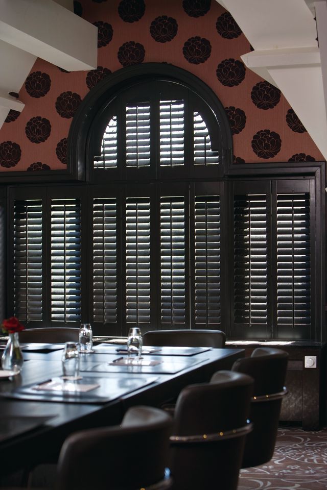 Dark wooden shutters cover an arched window in a formal conference room with patterned wallpaper and leather chairs.