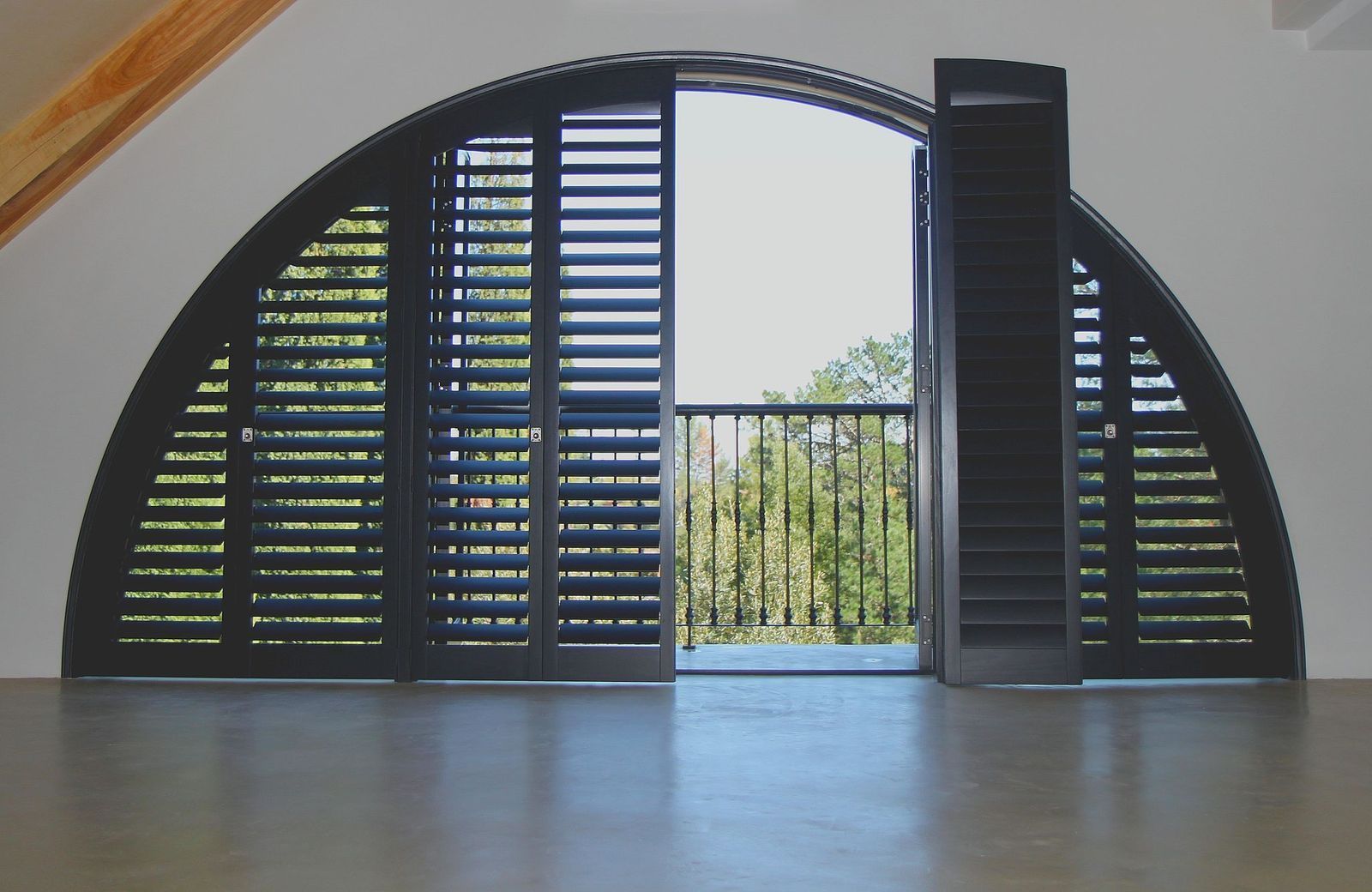 A semi-circular window with dark, slatted shutters, one of which is open to reveal a balcony with a metal railing.