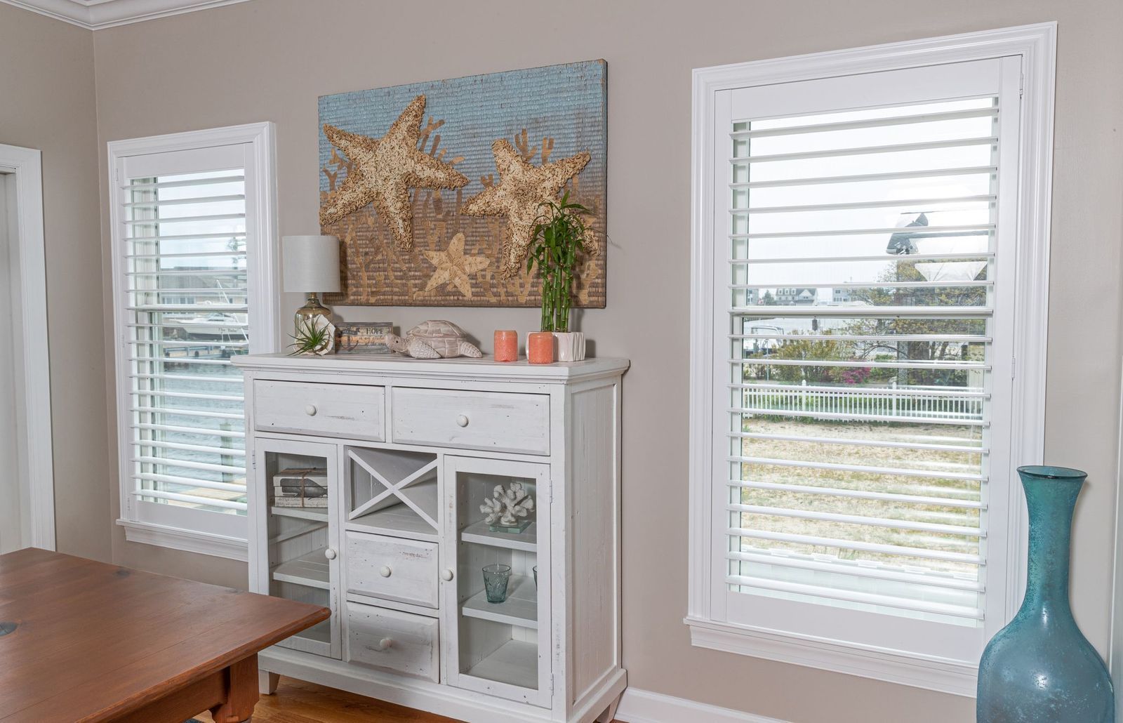 A white wooden buffet cabinet stands between two windows with white plantation shutters under a beach-themed artwork.