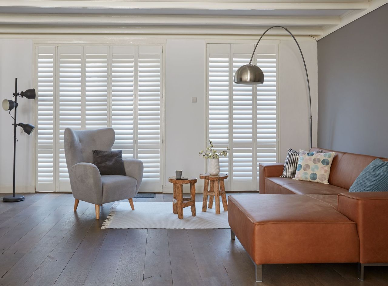 A living room features a grey armchair, a tan leather sofa with pillows, two wooden stools, and white window shutters.