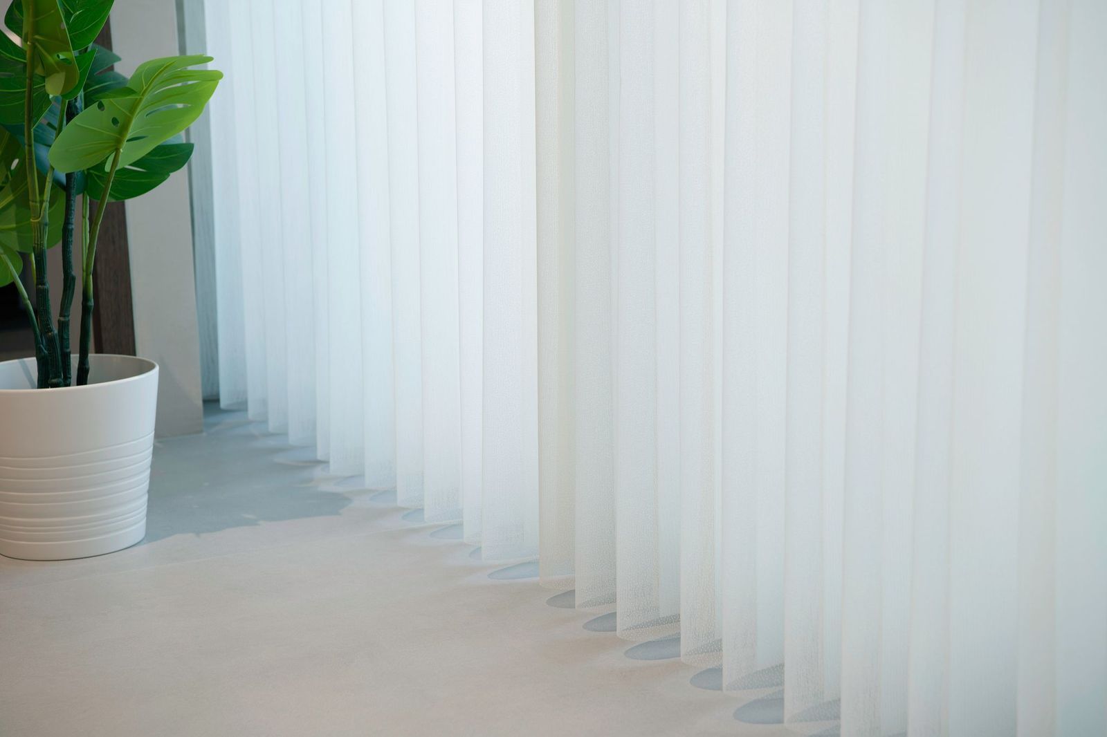 A white potted plant stands next to white, floor-length vertical blinds covering a window.