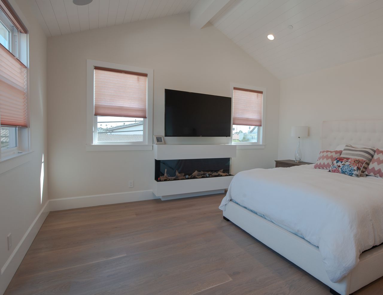 A bright bedroom features a king bed with patterned pillows, a wall-mounted TV above a fireplace, and light wood floors.