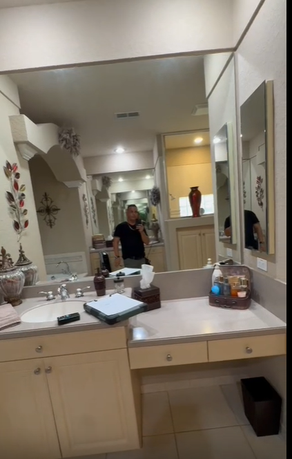 A person stands in front of a bathroom mirror, reflecting a vanity with a sink, drawers, and decor in a beige room.