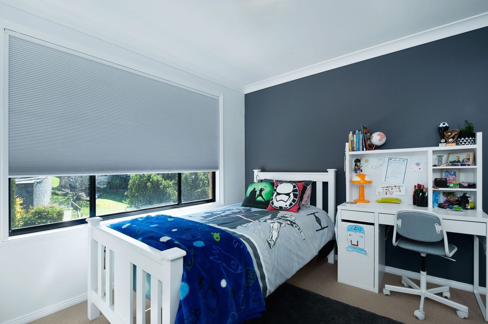 A child's bedroom featuring a white bed, a desk with a chair, and a large window with gray honeycomb blinds.