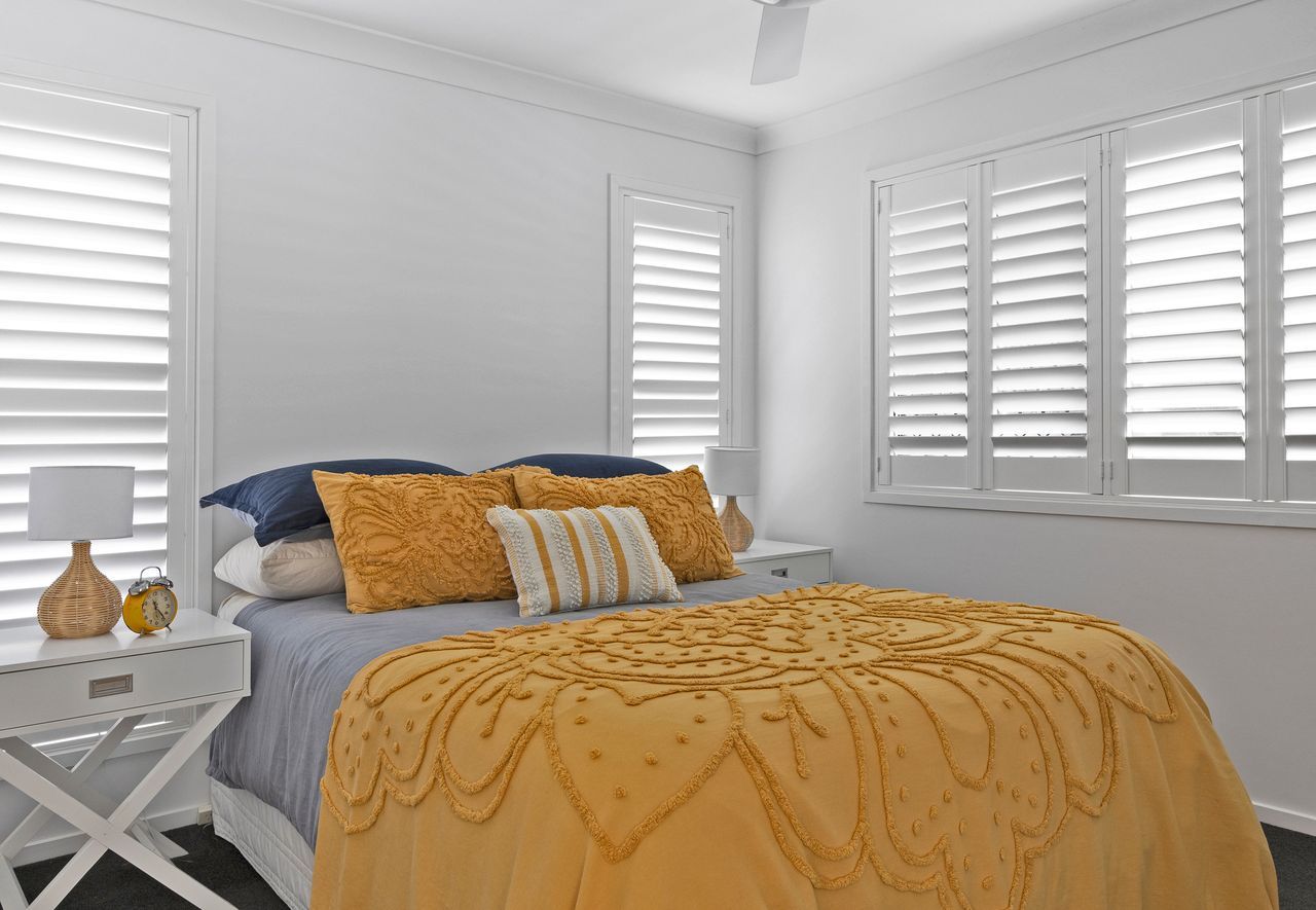 A bedroom with a yellow textured comforter and navy pillows on the bed, flanked by white nightstands and window shutters.