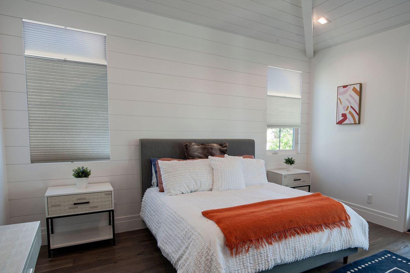 Modern bedroom with white shiplap walls, a gray bed with orange throw, and two nightstands with windows above them.