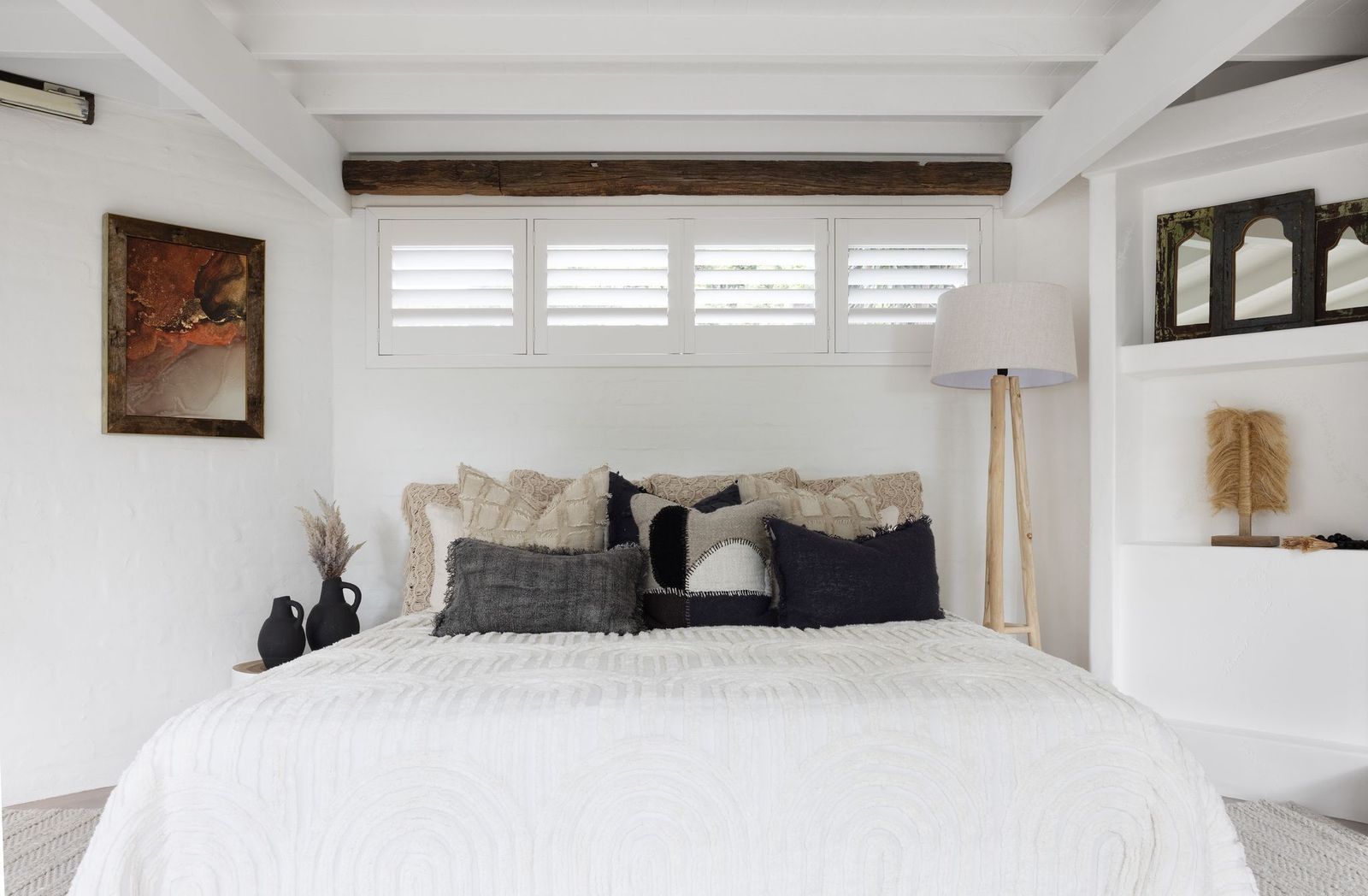 A cozy bedroom with a white bed, neutral-toned pillows, plantation shutters, exposed ceiling beams, and a floor lamp.
