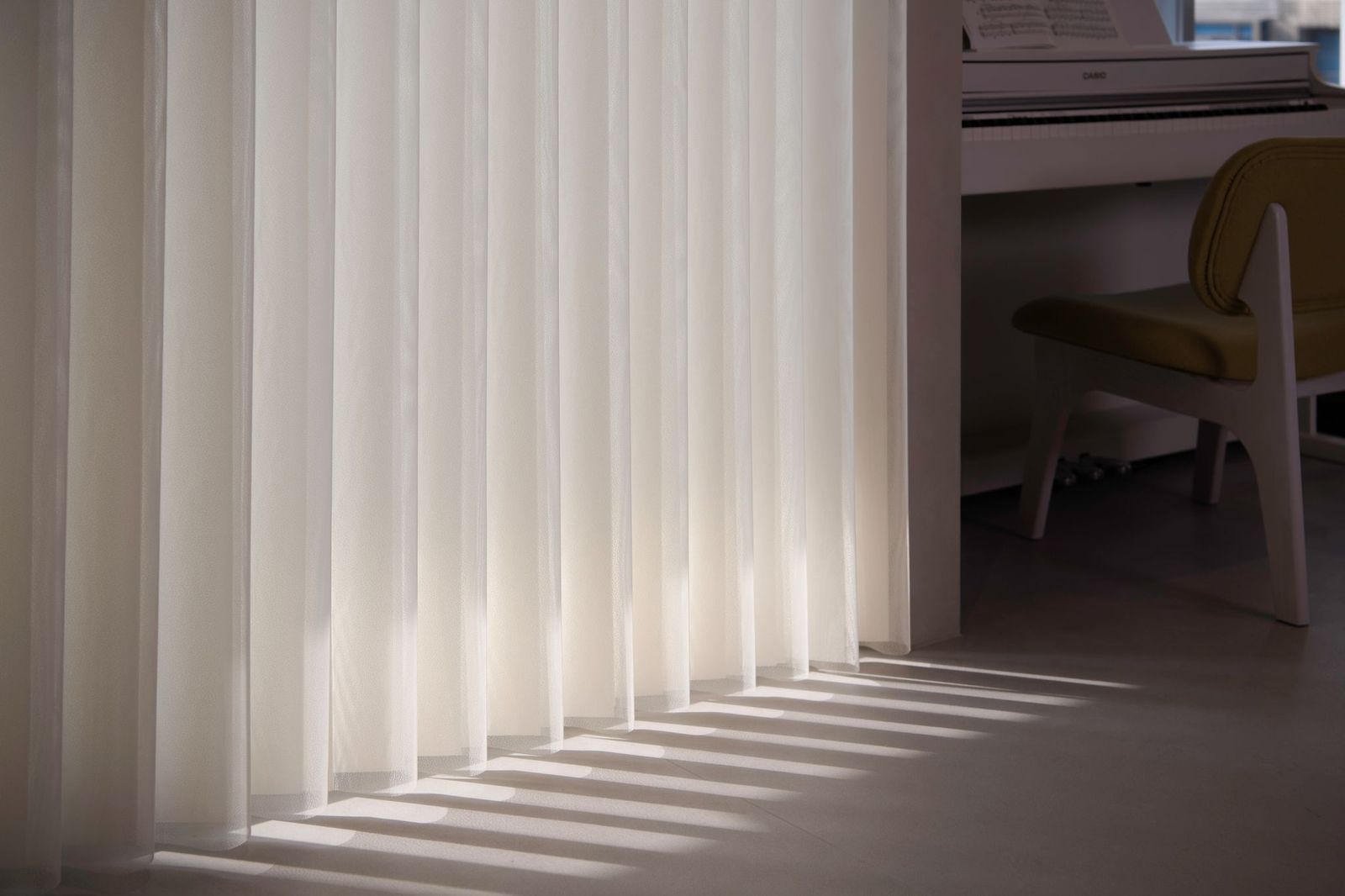 White vertical blinds filter sunlight, casting rhythmic, parallel shadows across a light-colored floor near a chair.