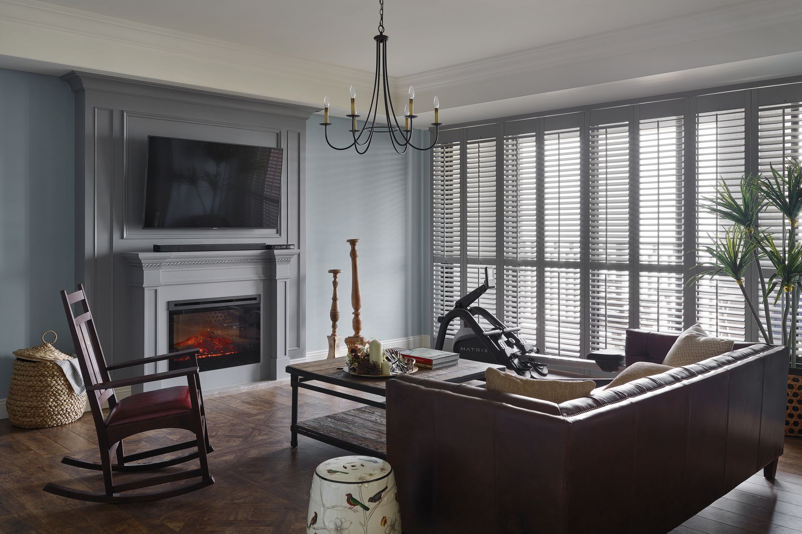 A cozy living room with a fireplace, TV, dark leather sofa, rocking chair, and large windows with plantation shutters.