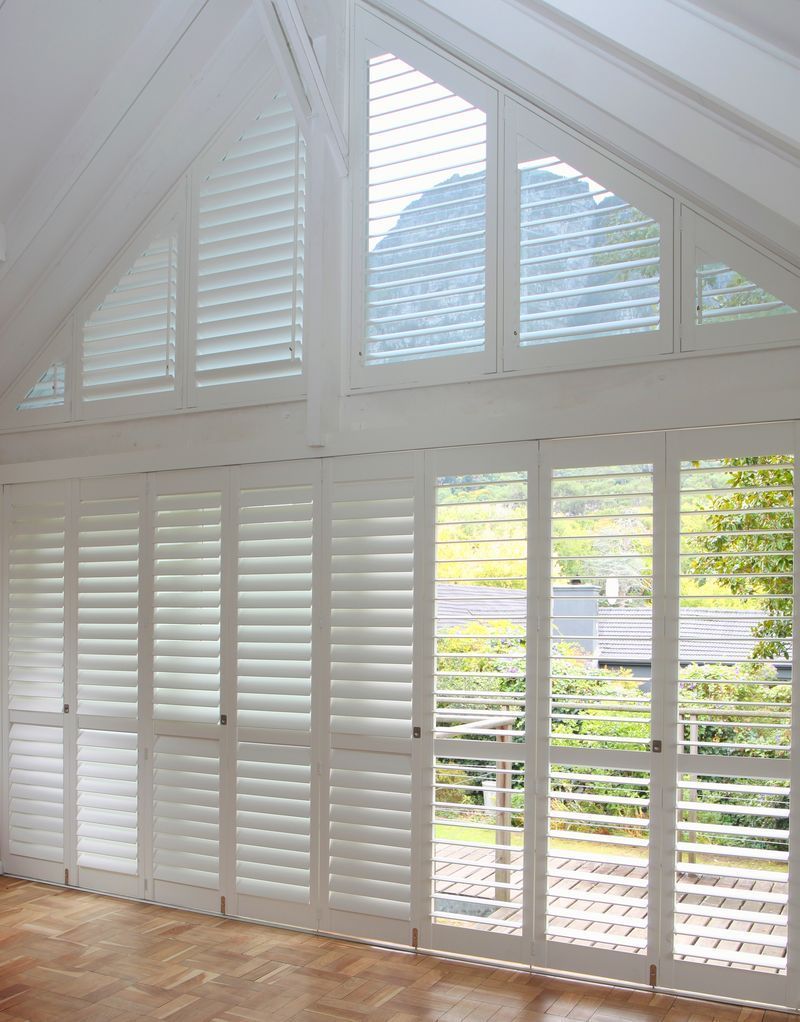 White wooden plantation shutters covering a floor-to-ceiling glass wall and matching triangle-shaped windows.