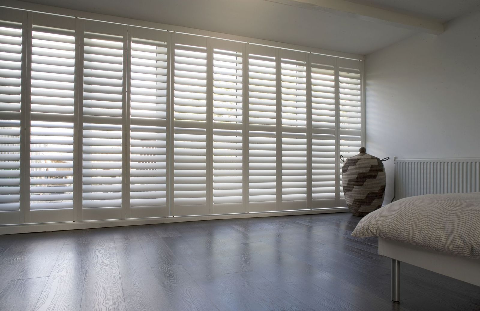 White floor-to-ceiling plantation shutters cover a wall of windows in a bedroom with dark wood flooring.