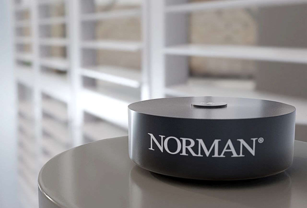 A black Norman-branded circular device sits on a surface, with blurred white window shutters in the background.